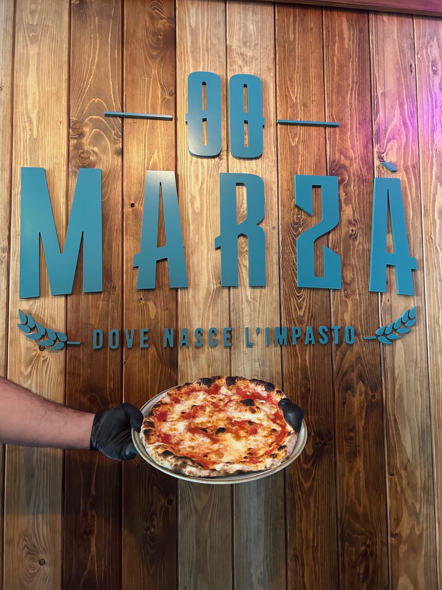 Pizza appena sfornata di Marzà davanti alla parete con il logo MARZÀ e la scritta Dove nasce l'impasto